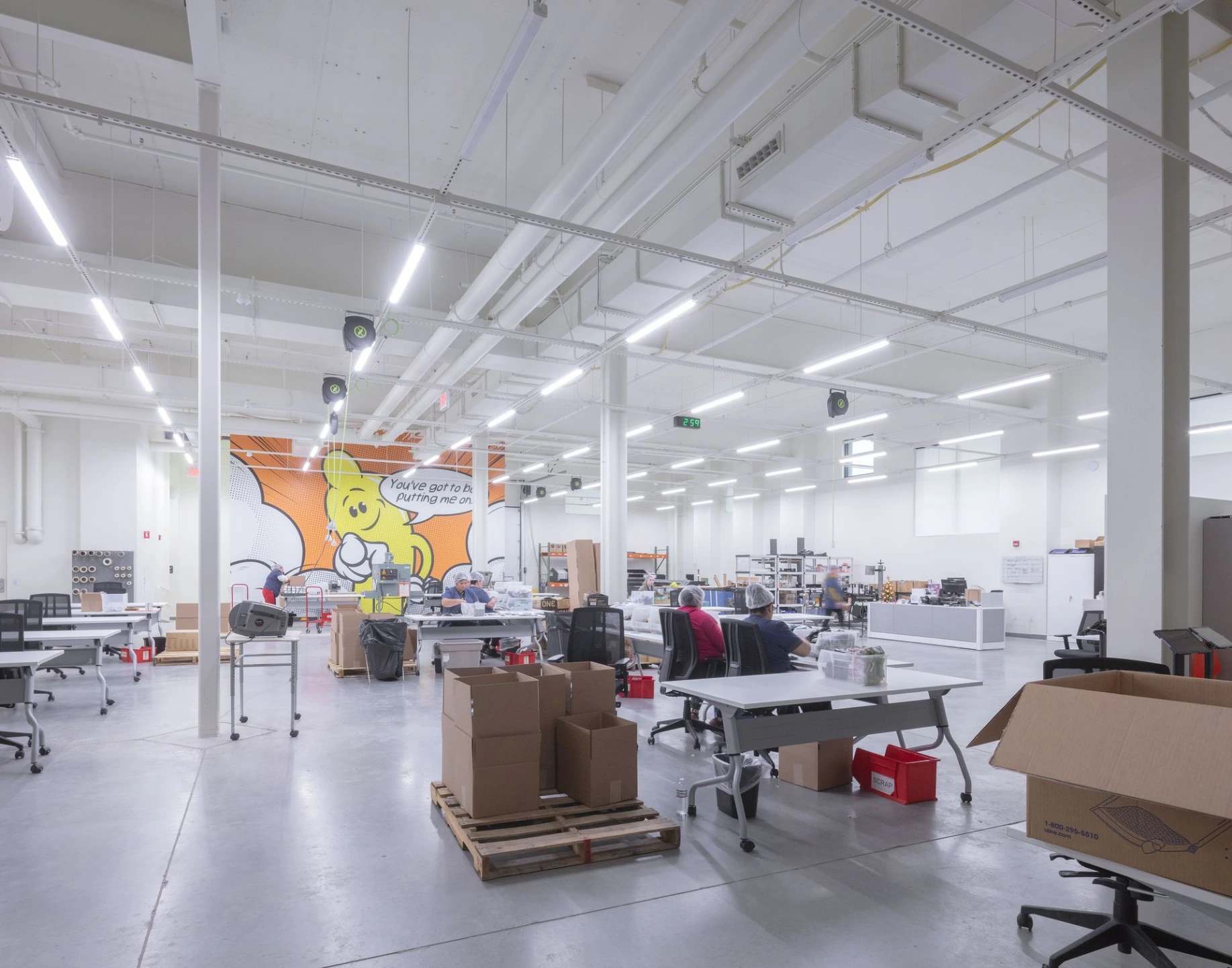 An active, brightly lit open-plan manufacturing and fulfillment workspace with employees at workbenches and a colorful geometric mural on the back wall.
