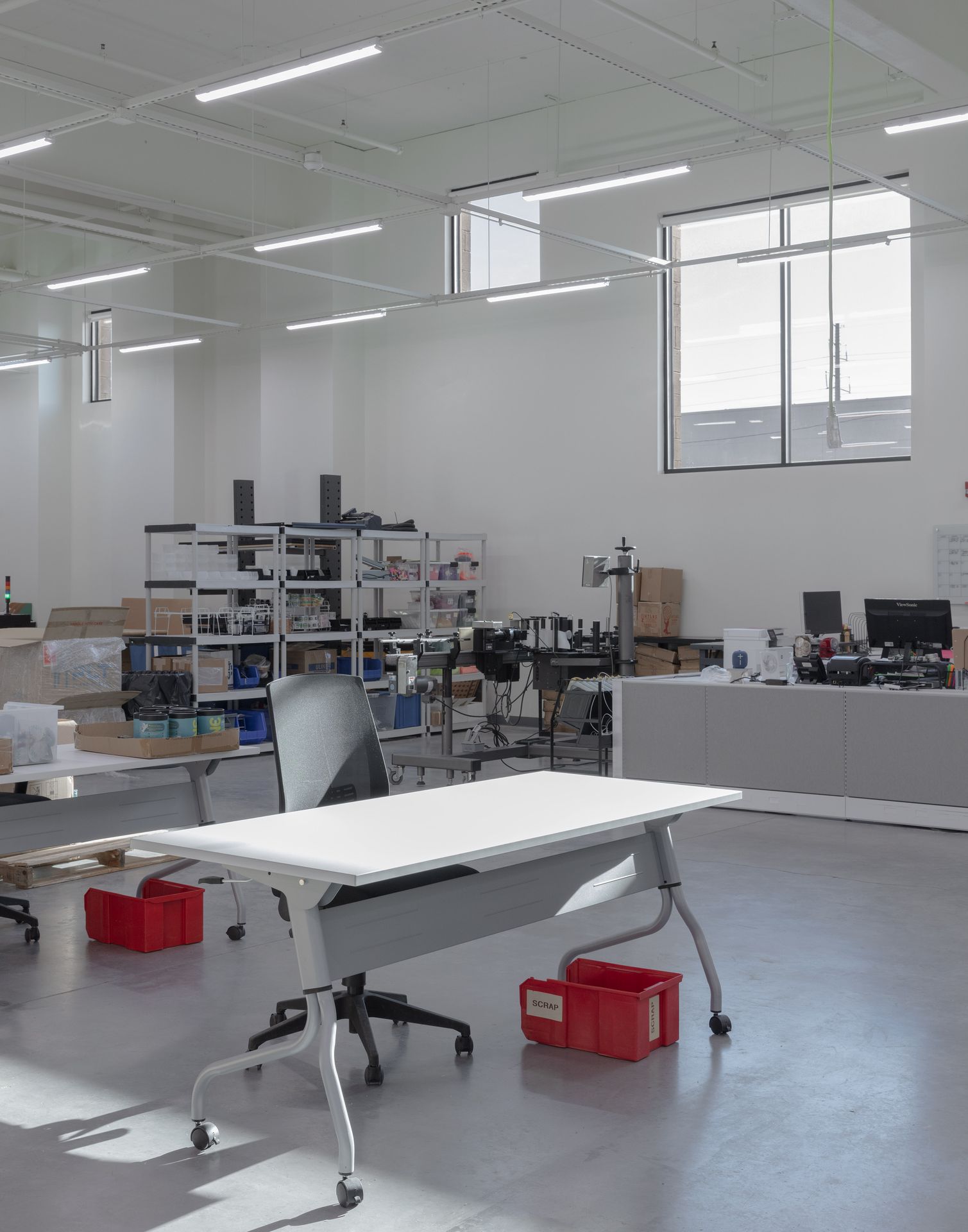 An open-concept corporate office workspace featuring a long white communal desk, task seating, and large windows bringing in natural light.