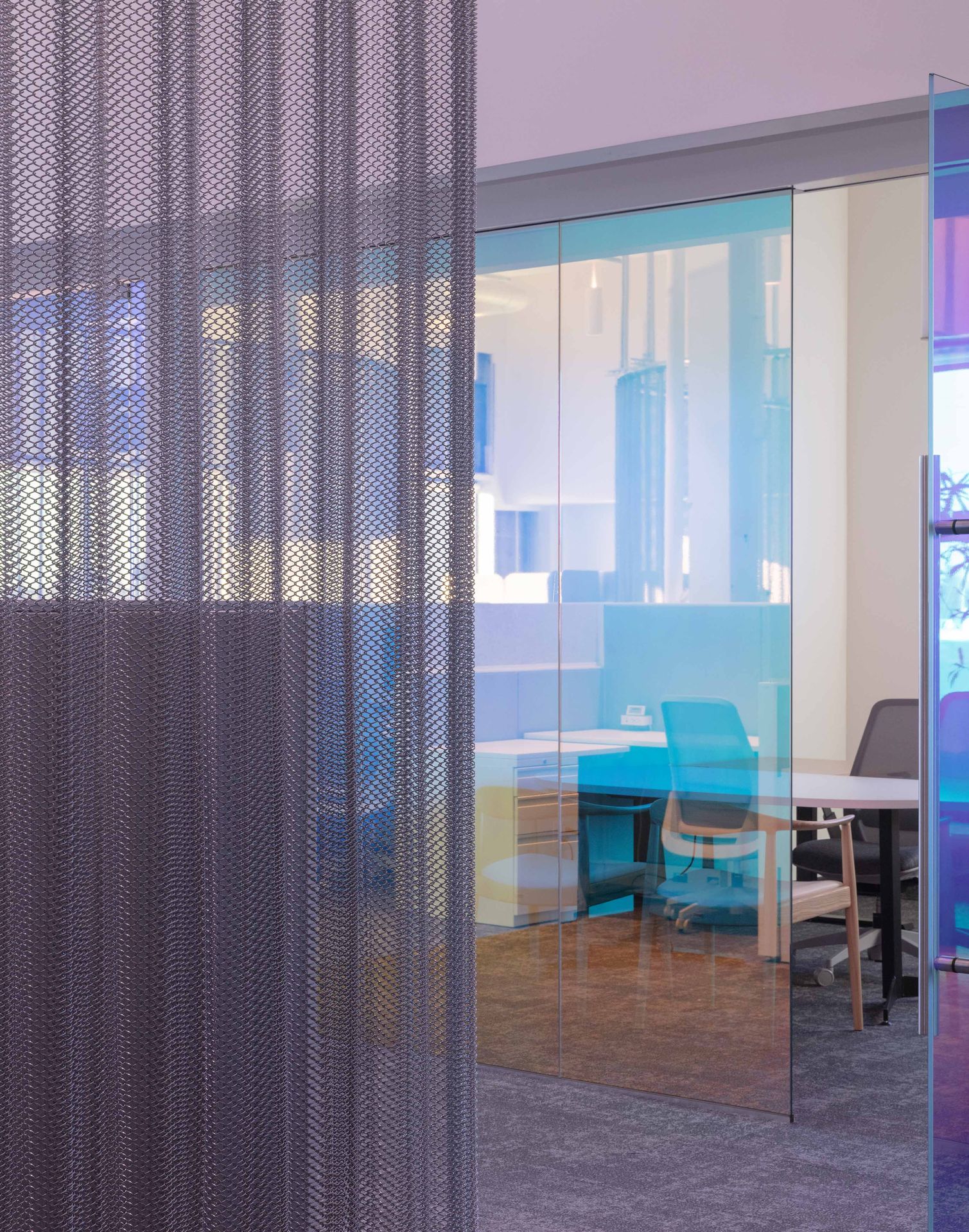 View looking through a metallic blue chainlink or mesh partition wall separating bright office corridors.