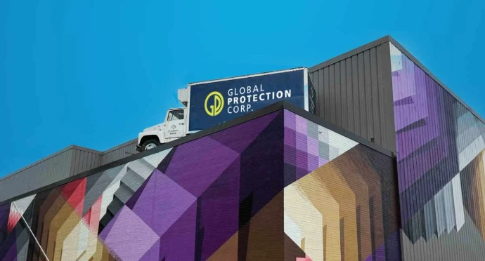 Global Protection Corp. Building A low-angle exterior shot of a modern building featuring a vibrant, multi-colored geometric mural in shades of purple, gold, and red. A white delivery truck with the "Global Protection Corp." logo is parked on the roof or a high elevated platform against a clear blue sky.