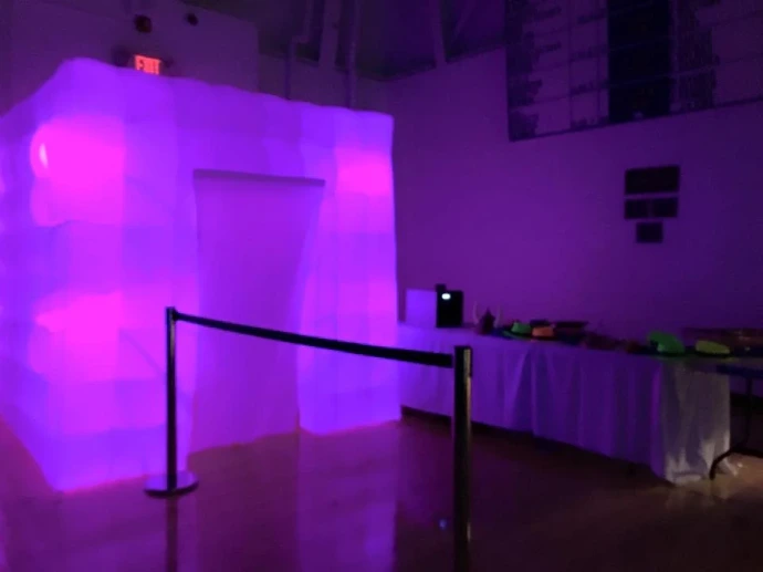 A large, glowing purple inflatable LED photo booth cube set up at an event next to a table of props and a velvet rope barrier.