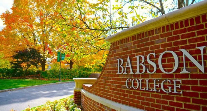 A brick entrance sign for Babson College in Wellesley, Massachusetts, surrounded by vibrant orange and yellow autumn foliage and a paved driveway.